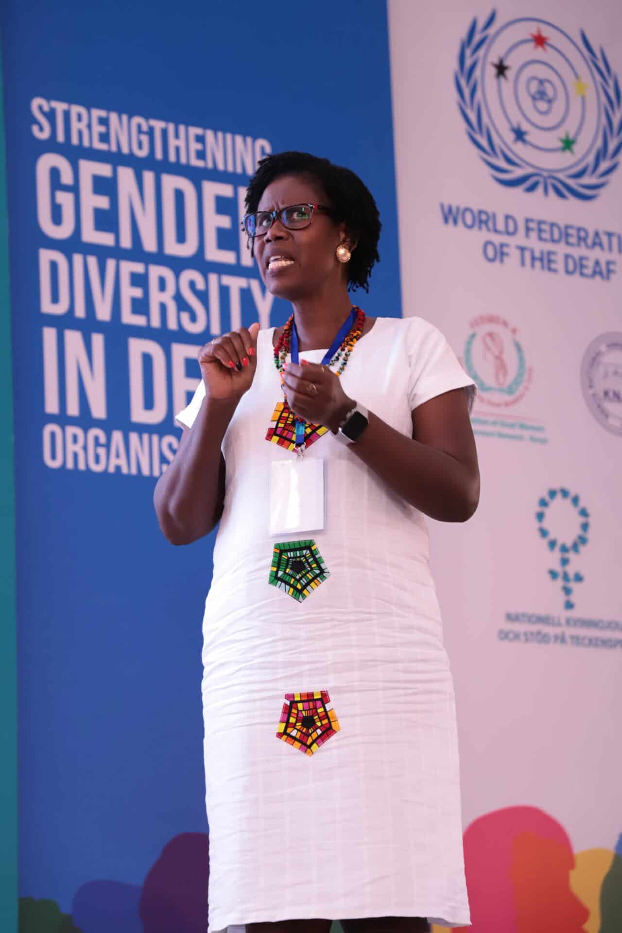 A woman in a white dress with colorful beadwork speaks on stage at a World Federation of the Deaf event focused on gender diversity in organizations.