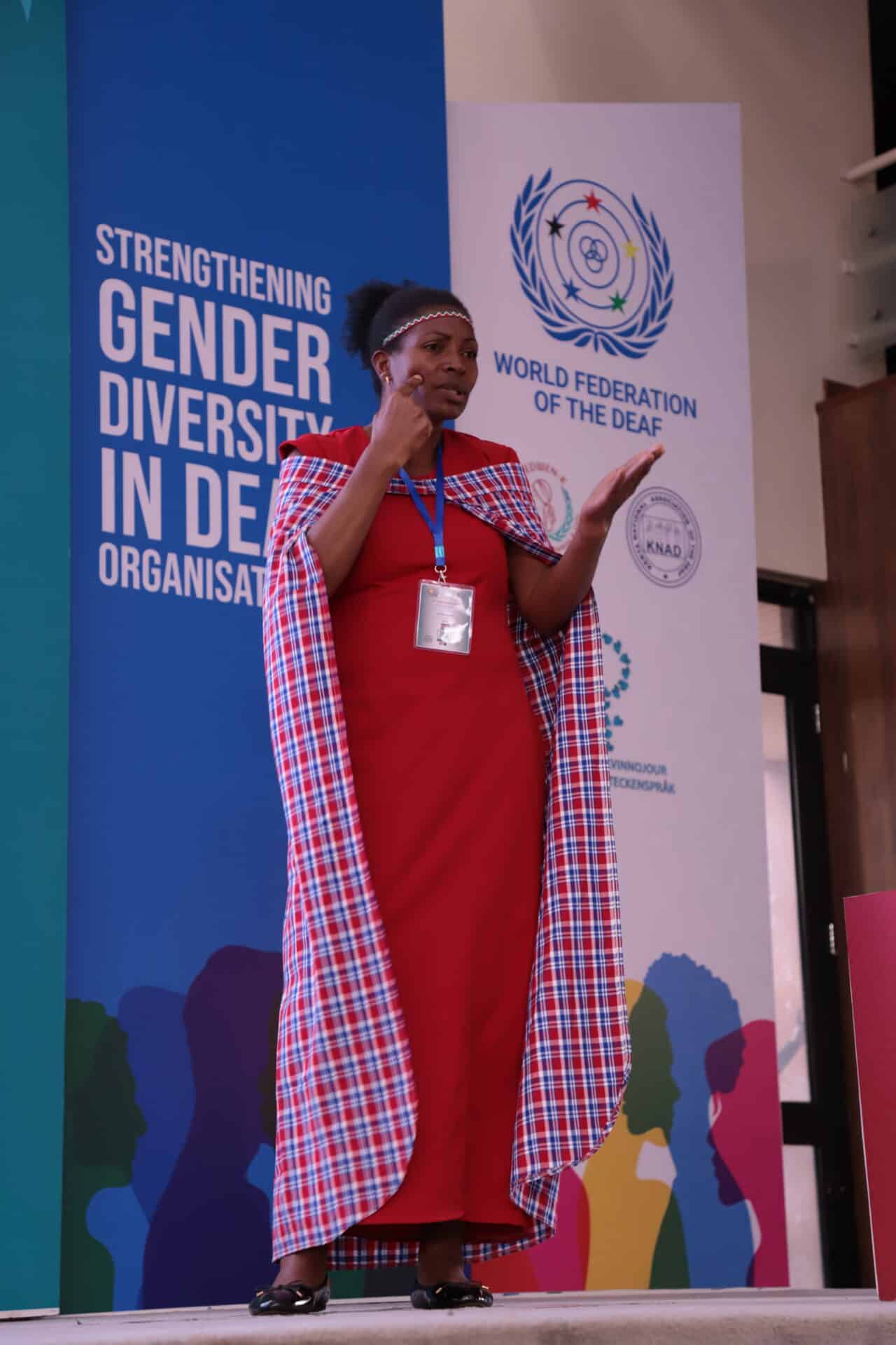 A woman in a red dress and patterned shawl uses sign language on stage at a World Federation of the Deaf event focused on strengthening gender diversity.
