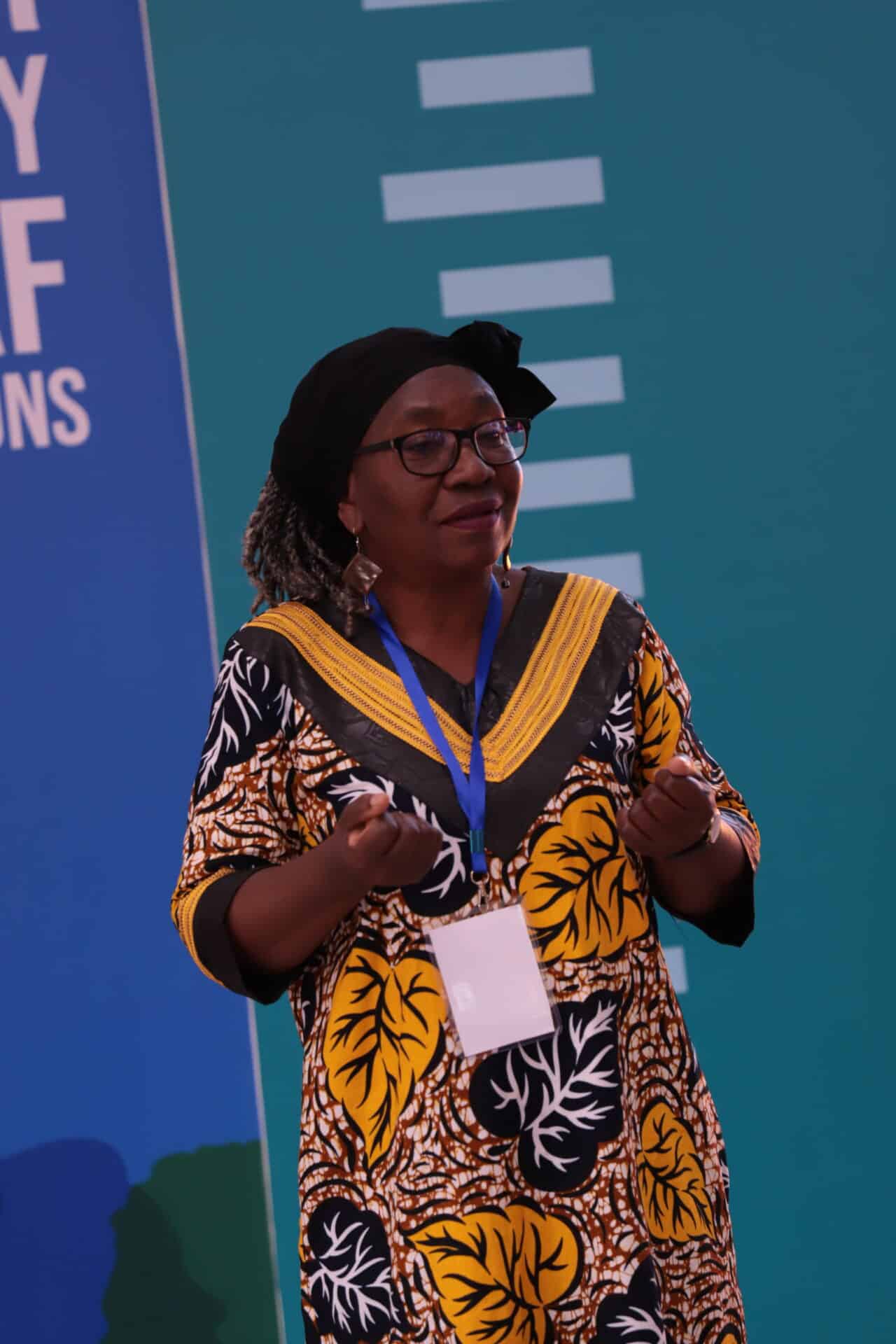 A woman wearing glasses and a colorful dress speaks at an event, wearing a blue lanyard and name badge, with a teal background behind her.