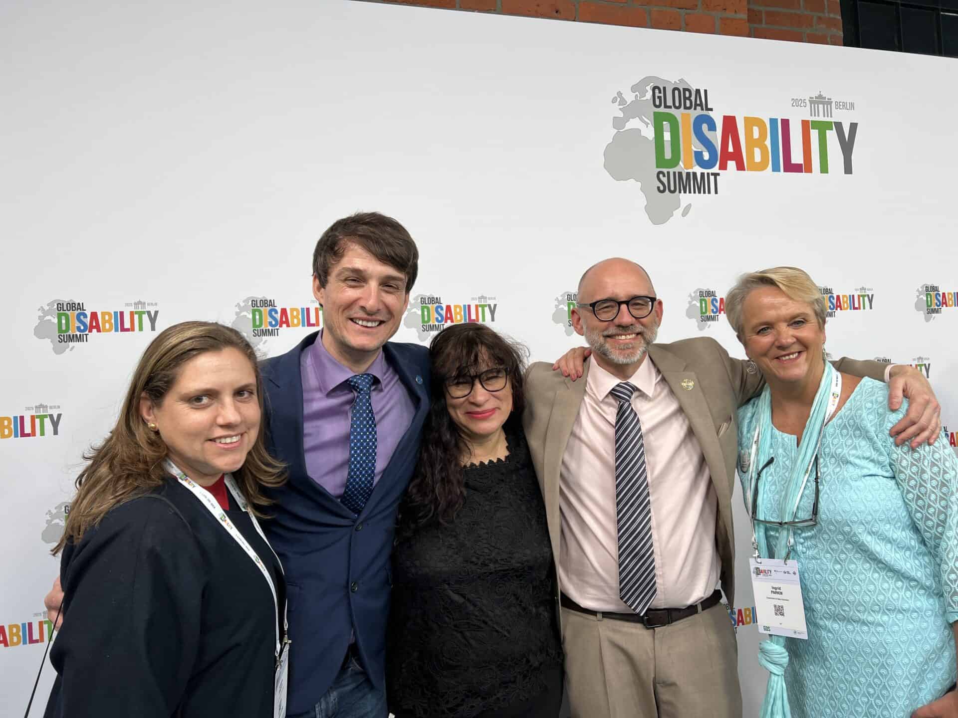 Five people standing together and smiling in front of a Global Disability Summit backdrop.