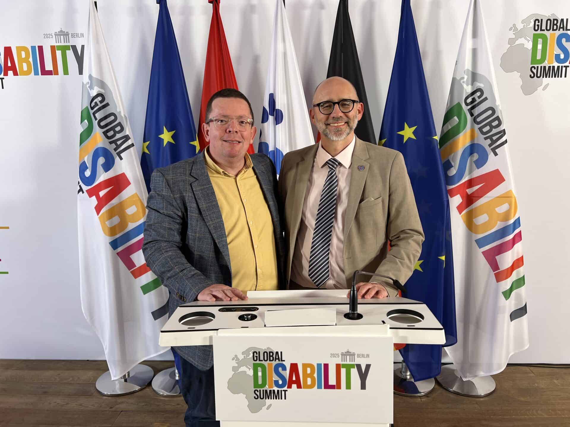 Two men stand together smiling in front of a podium labeled "Global Disability Summit," with various international flags in the background.
