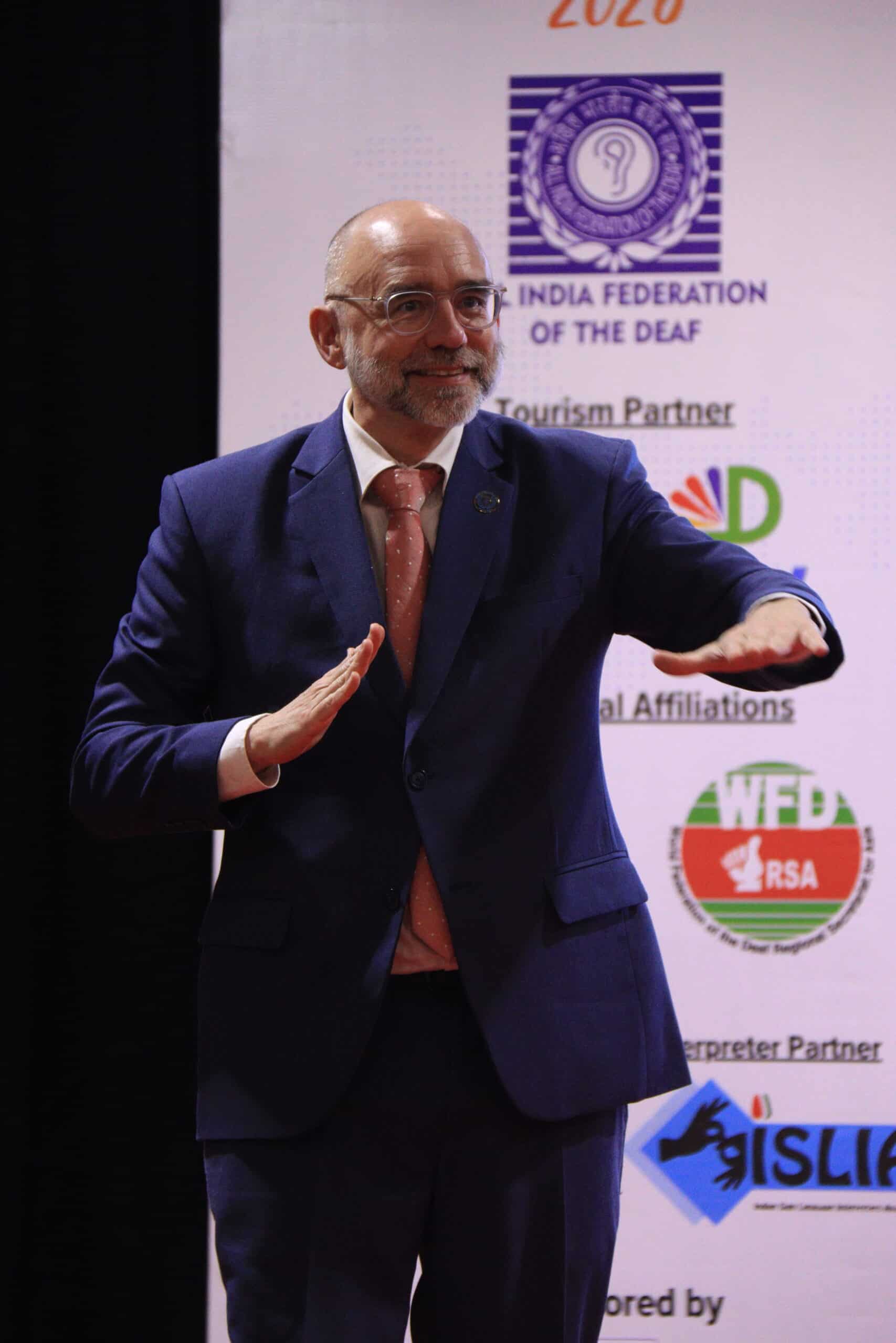 A man in a blue suit stands in front of a banner with logos, gesturing with both hands at waist height.