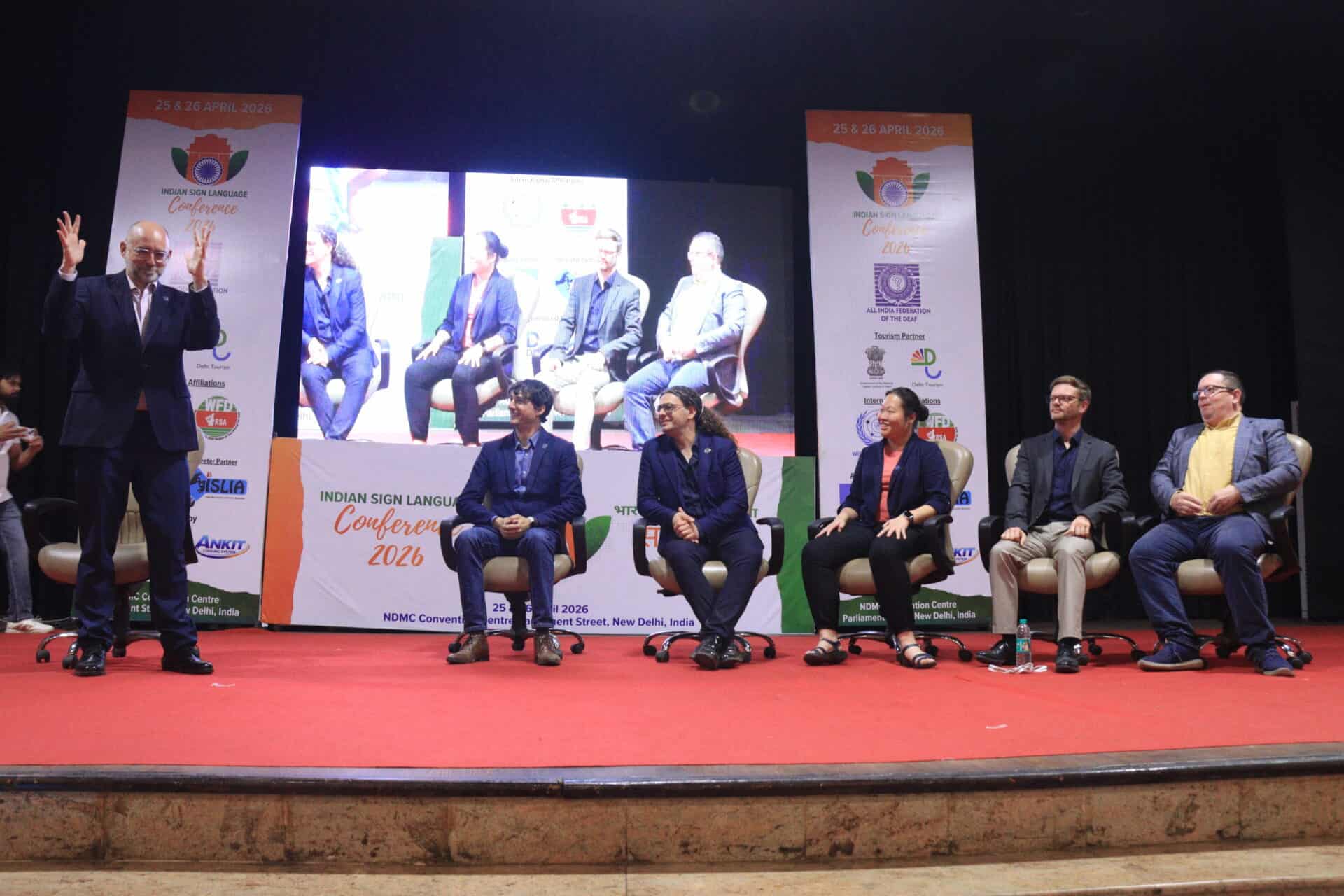 Six people are seated on a stage while one person stands to the left, gesturing with raised hands. Conference banners and a screen are displayed in the background.
