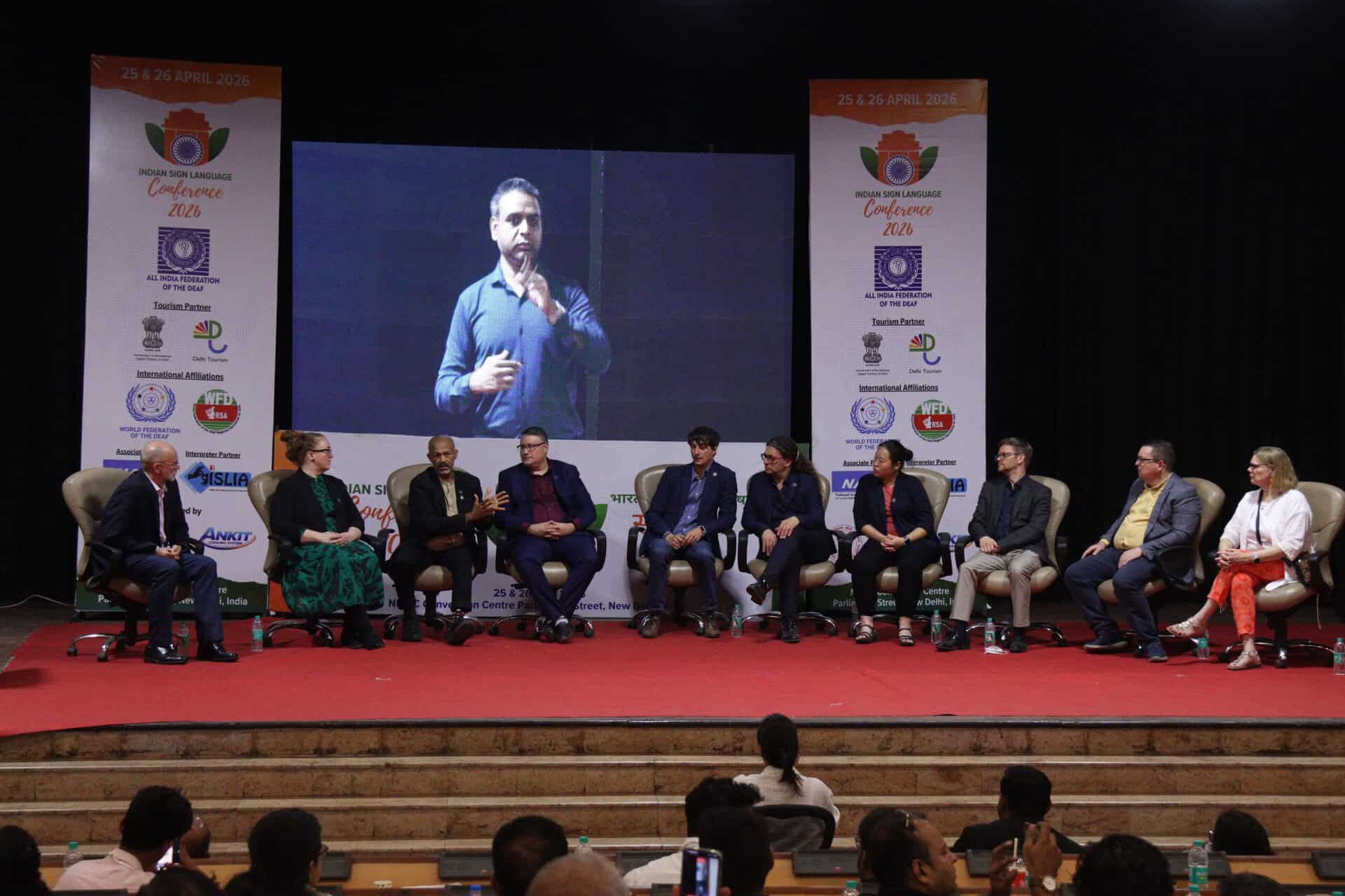A panel of ten people sits on stage, with one person standing and speaking into a microphone in front of a large screen, during a conference event.