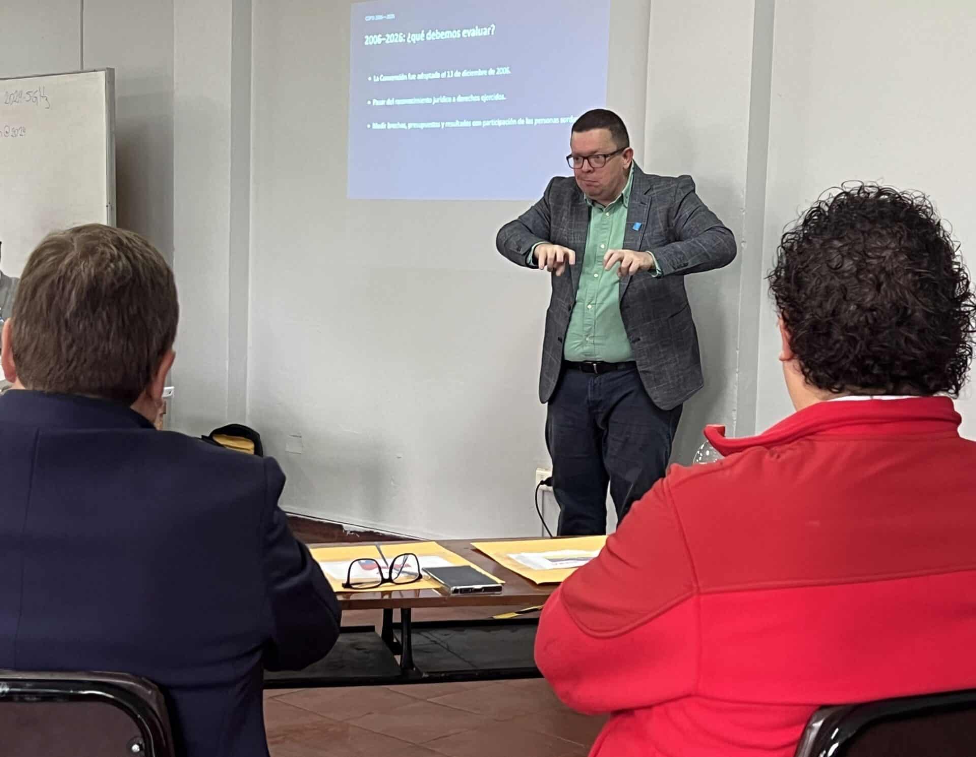A man in a green shirt and grey blazer gestures with his hands whilst standing in front of a projector screen showing a presentation.