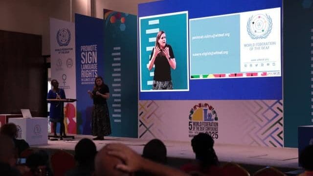 A speaker presents at a conference on sign language rights, with a sign language interpreter and informational slides displayed on a large screen.