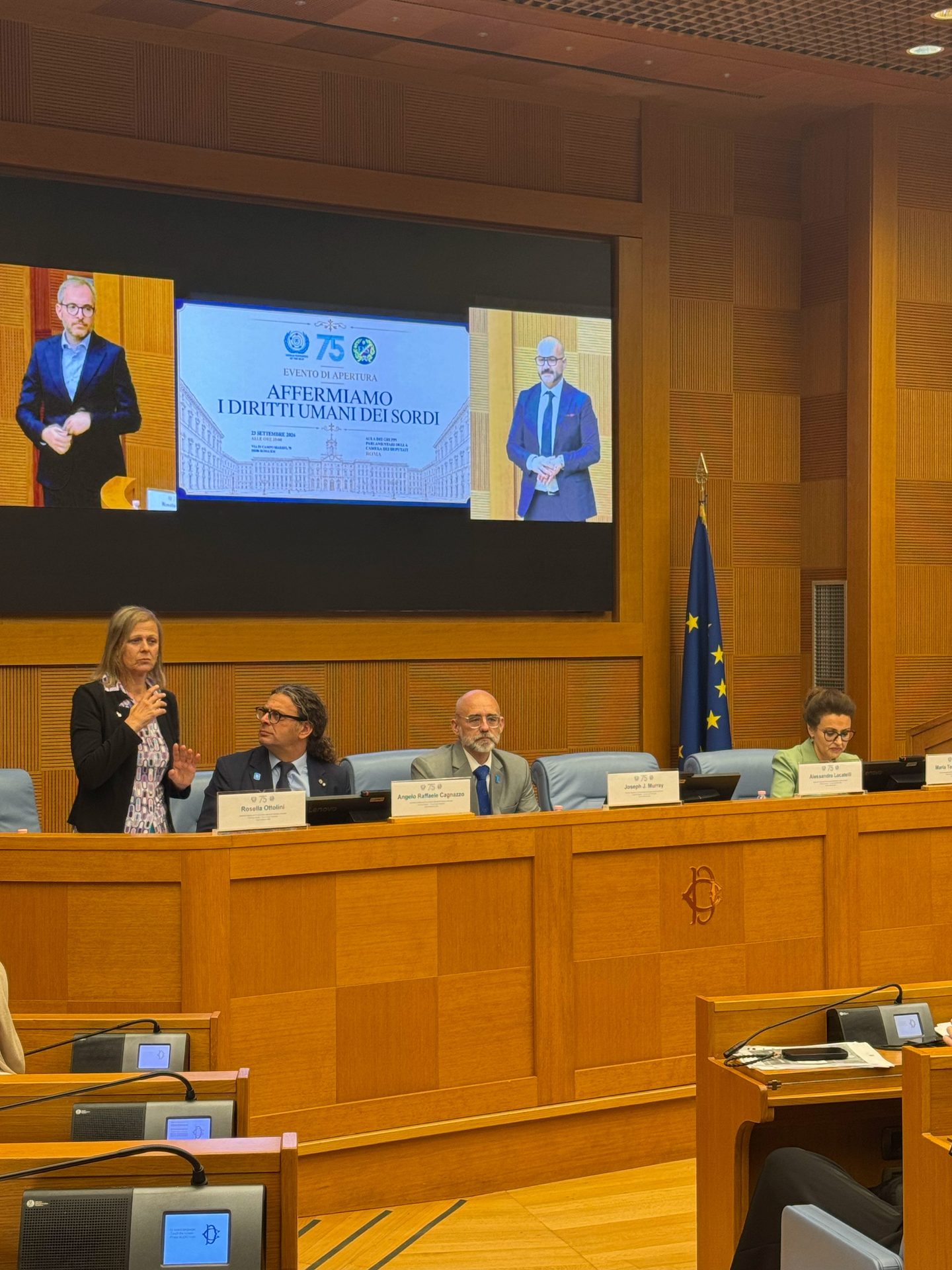 Four people sit at a conference table with microphones; a woman stands speaking. Behind them, a screen displays a message and two images of a man in a suit. An EU flag is visible.