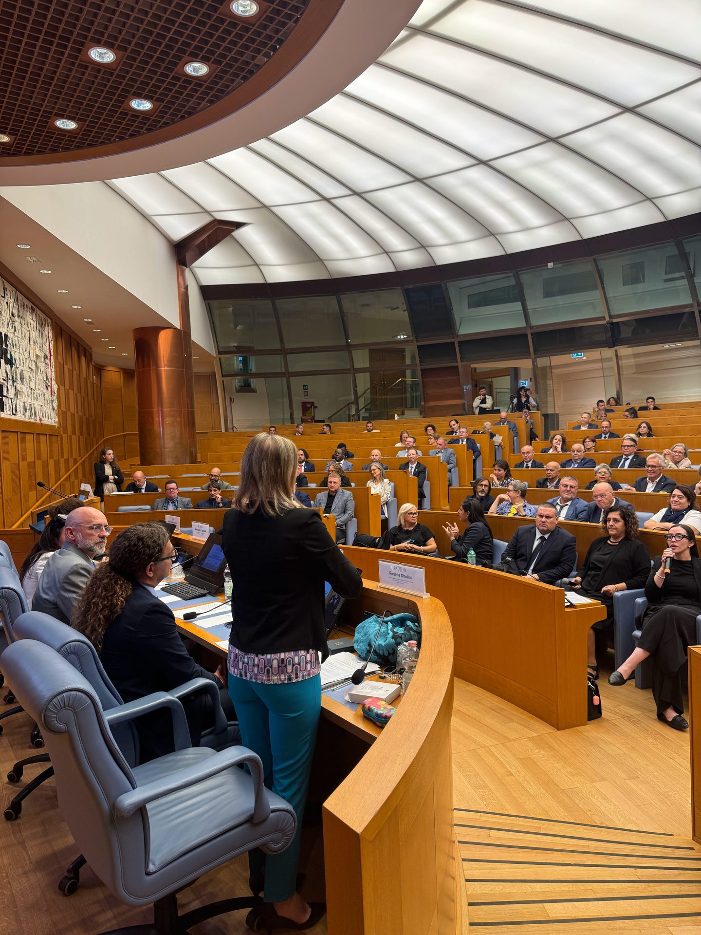 A woman stands at a lectern addressing a seated audience in a modern, wood-panelled conference room with tiered seating and a curved ceiling.