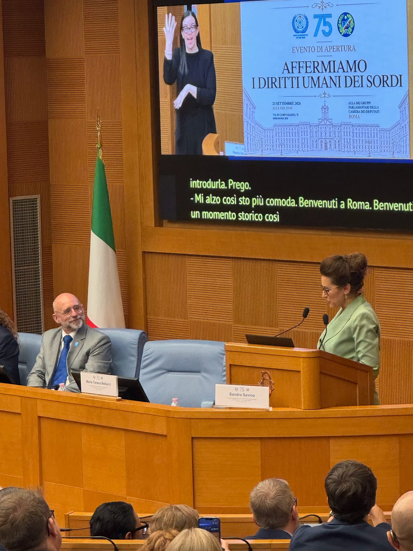 A woman speaks at a lectern in a conference room; behind her is a screen about affirming deaf human rights, with a man and Italian flag seated nearby.