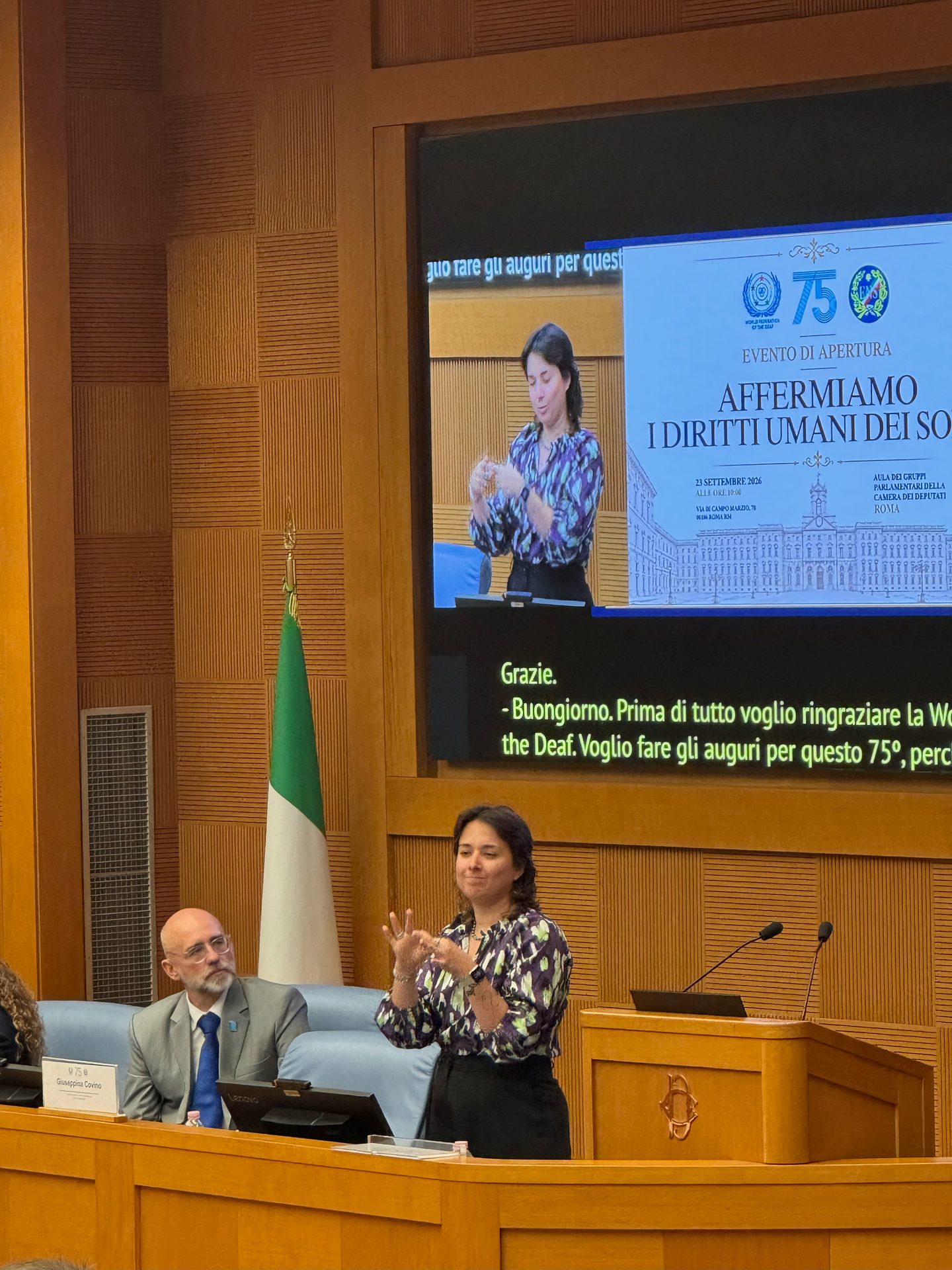 A woman signs at a lectern during a human rights event, while a man sits nearby. An Italian flag and presentation screen are visible in the background.