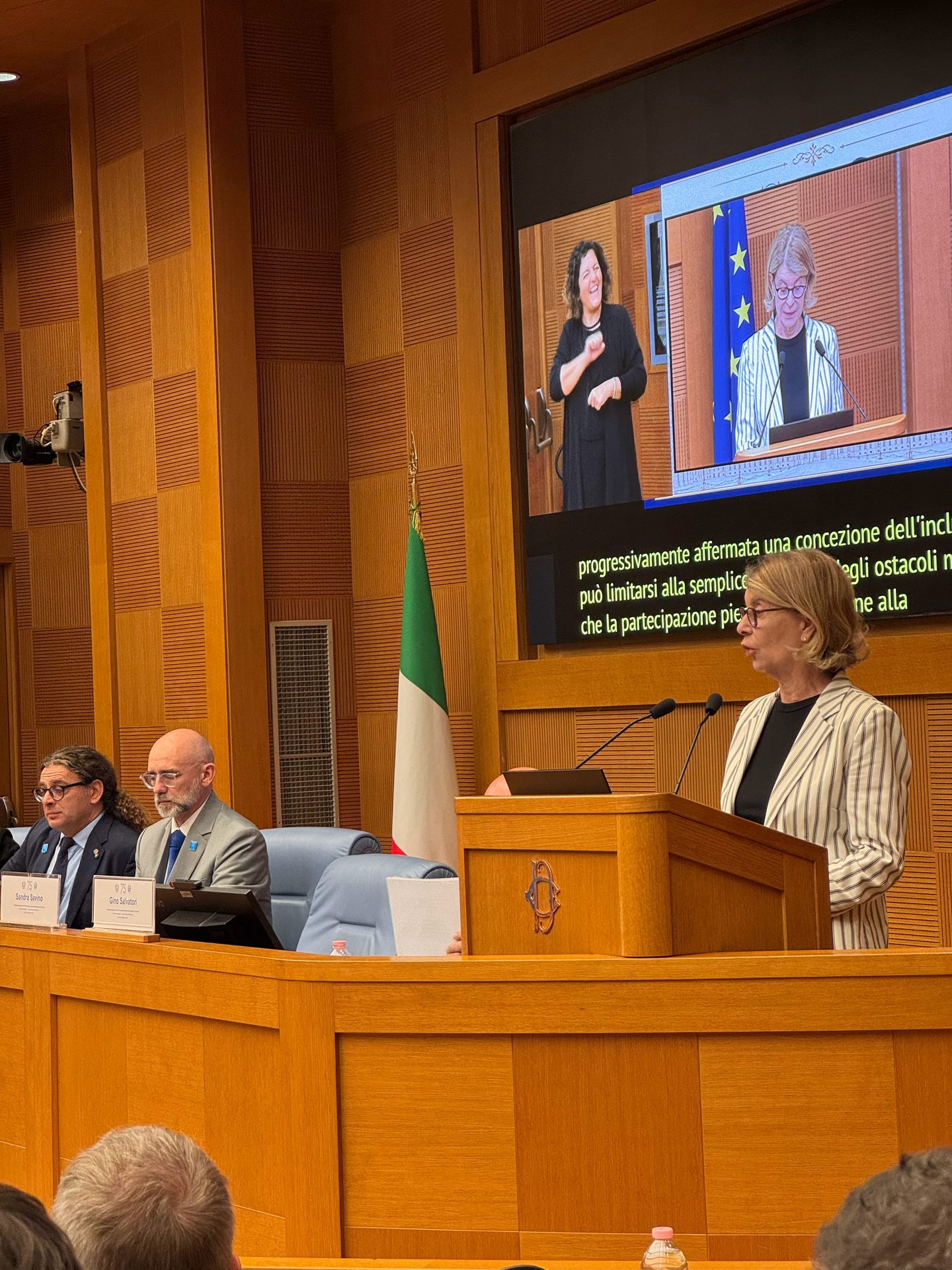 A woman speaks at a lectern in a wood-panelled room with two men seated nearby; an Italian flag and a screen showing a sign language interpreter are visible.