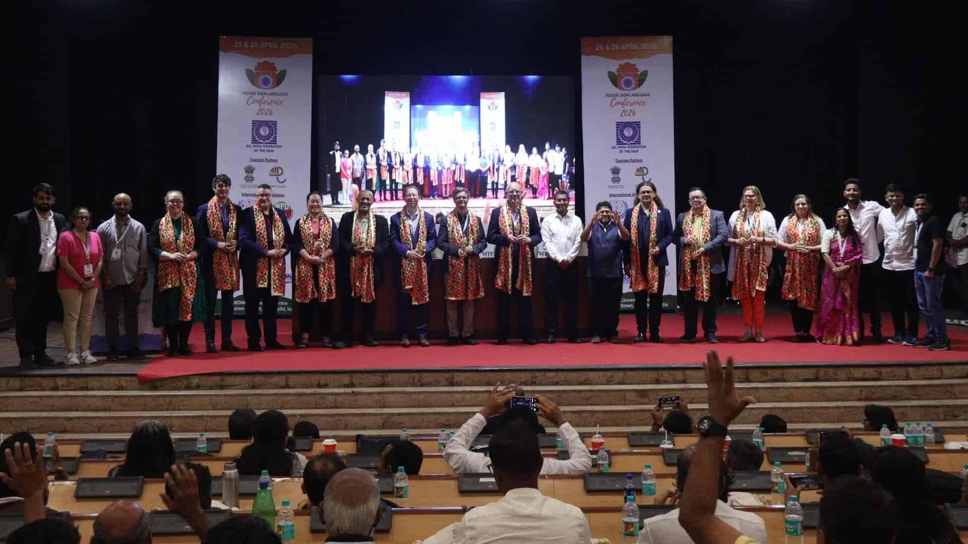 A group of people stand on stage at an indoor event, some wearing colourful sashes, with an audience seated in front of them taking photos.