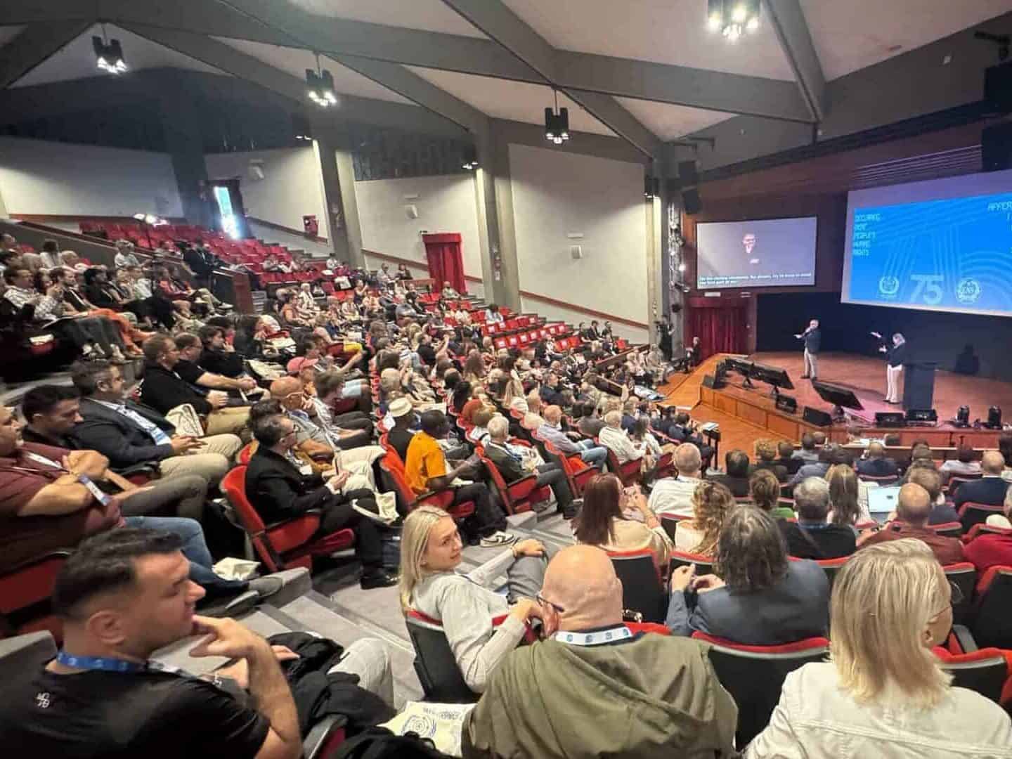 A large audience sits in a lecture theatre watching a presentation on a stage, with speakers in front of a screen displaying graphics and text.