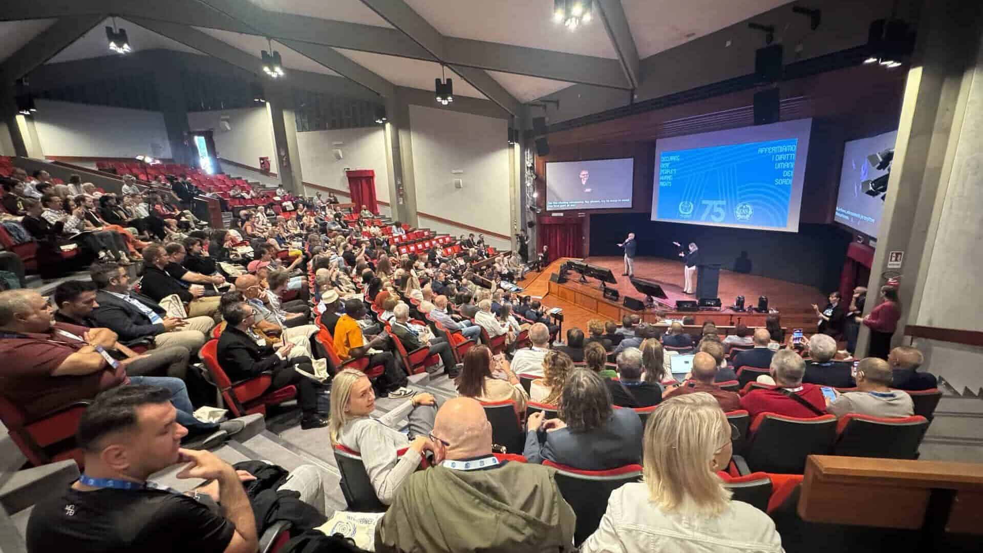 A large audience sits in a lecture theatre watching a presentation on a stage, with speakers in front of a screen displaying graphics and text.