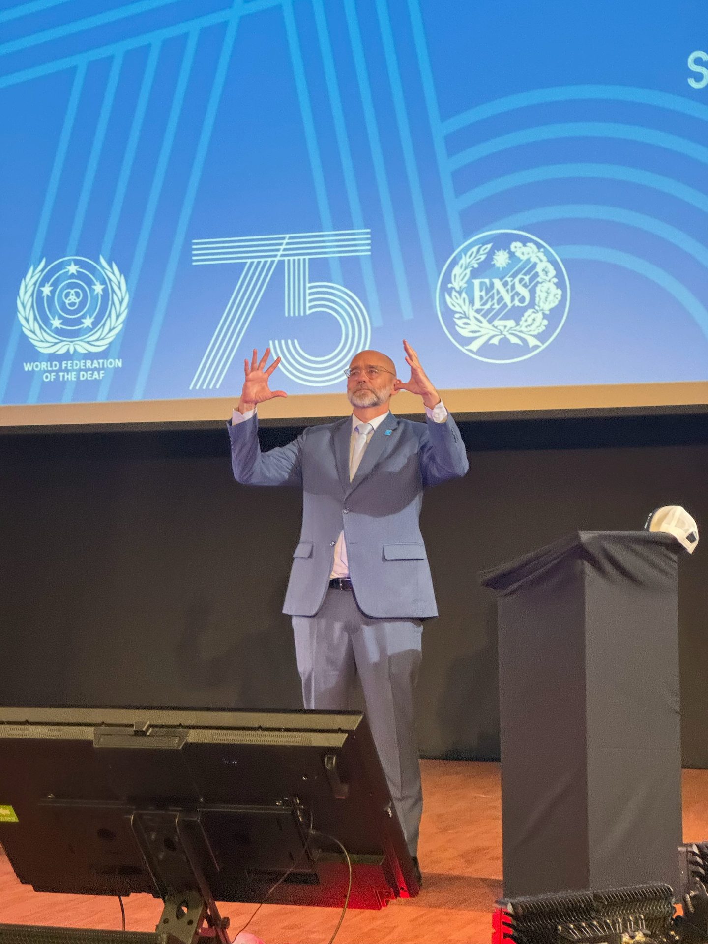 A man in a blue suit stands on stage gesturing with raised hands, with a blue background displaying “75” and organisational logos.