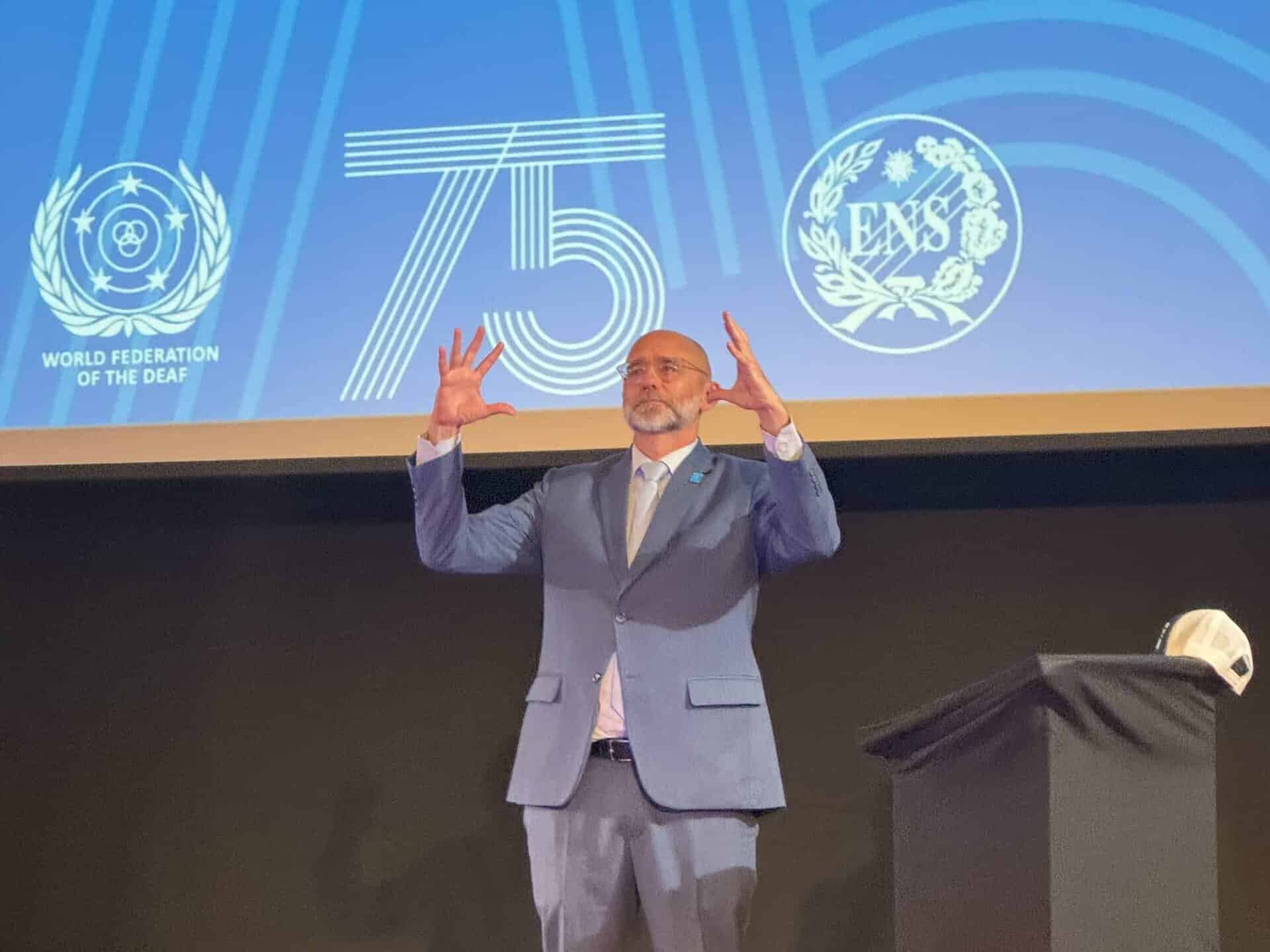 A man in a blue suit stands on stage gesturing with raised hands, with a blue background displaying “75” and organisational logos.