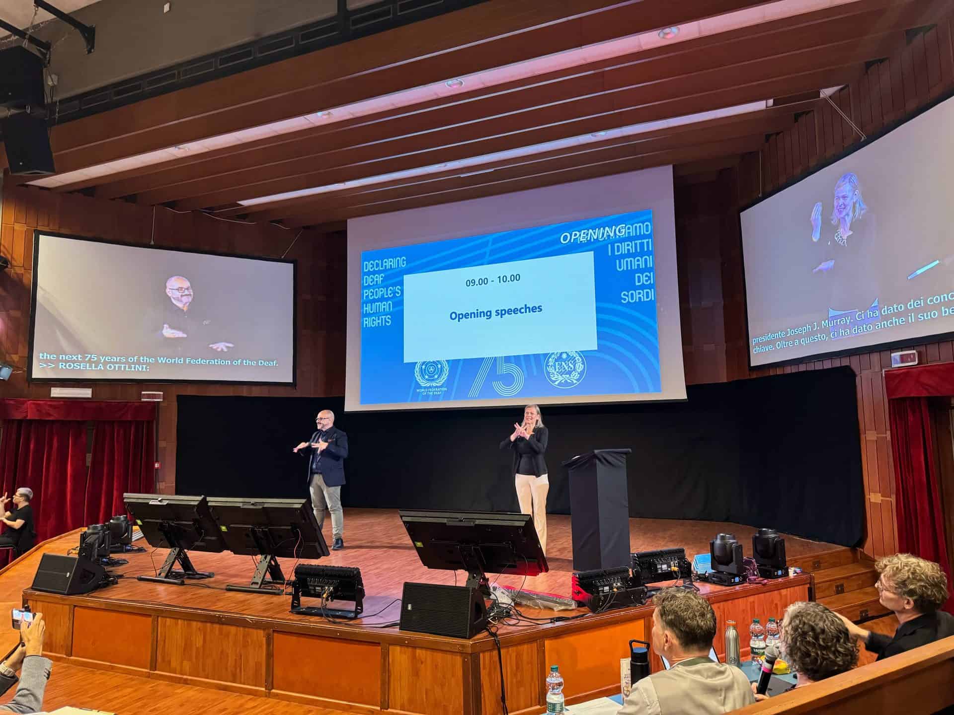 A conference stage with two sign language interpreters, a presentation screen displaying “Opening speeches”, and audience members seated in front.