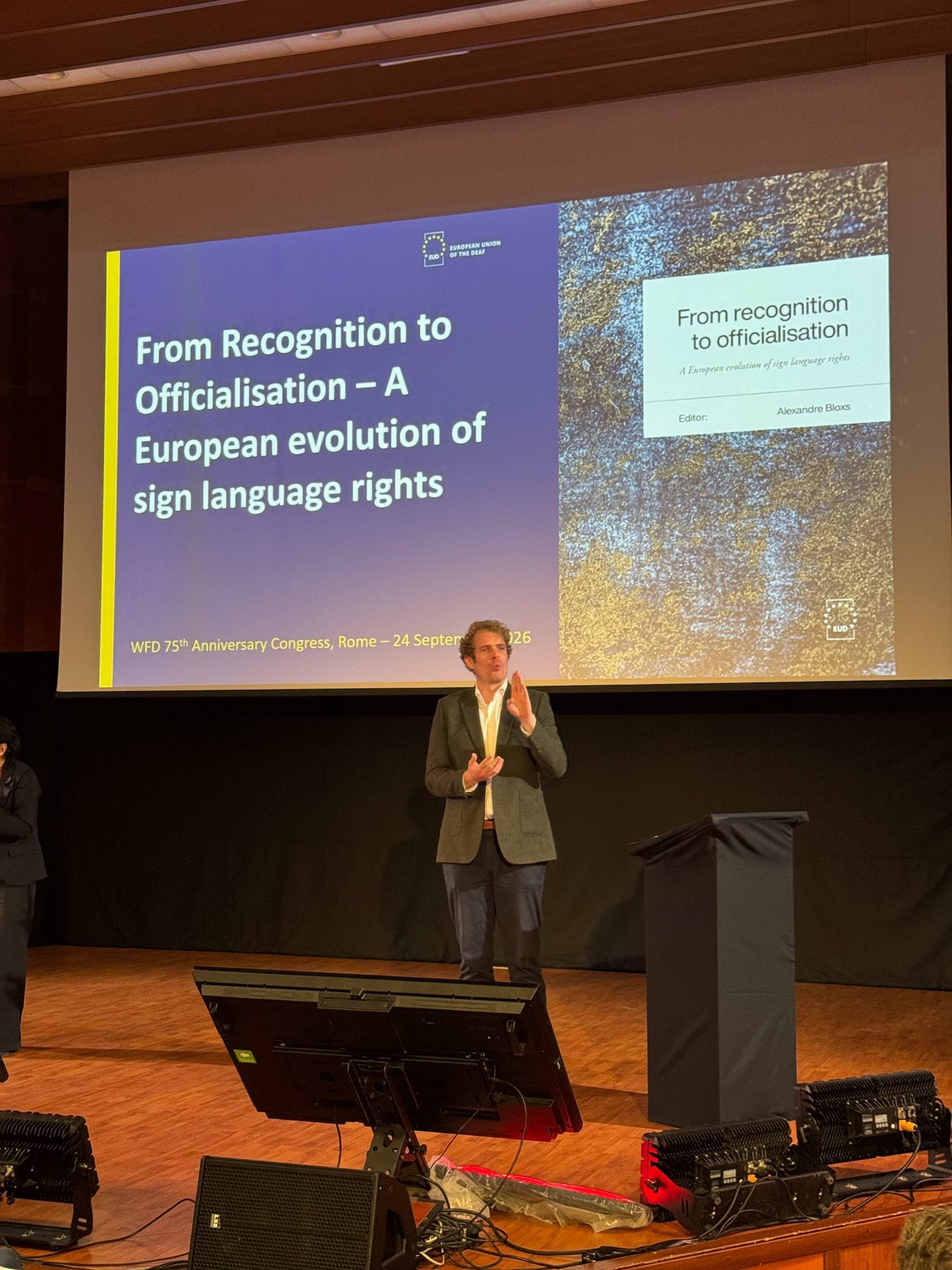 A person stands onstage speaking at a conference, with a presentation slide behind them titled “From Recognition to Officialisation – A European evolution of sign language rights.”.