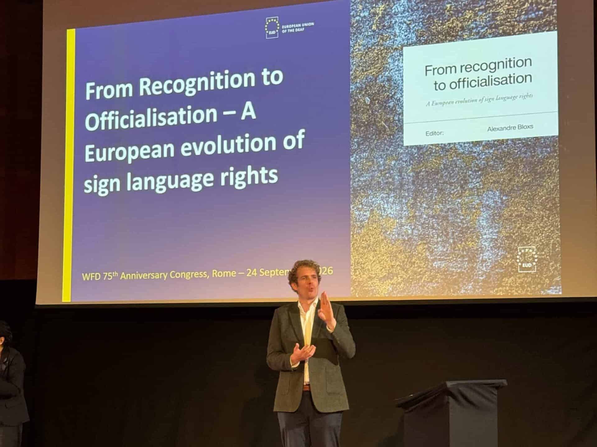 A person stands onstage speaking at a conference, with a presentation slide behind them titled “From Recognition to Officialisation – A European evolution of sign language rights.”.
