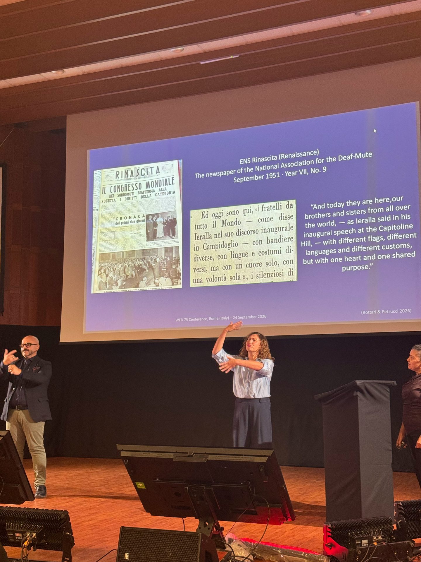 Three presenters stand on stage in front of a large screen displaying a historical newspaper and text about the National Association for the Deaf-Mute from September 1951.