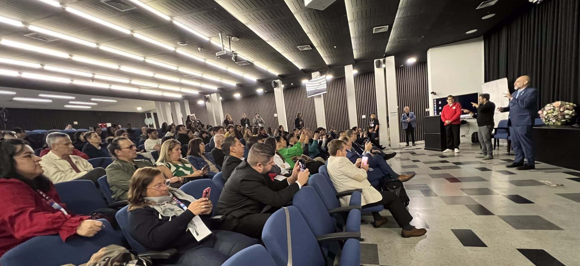 An audience seated in an auditorium watches a man presenting at the front of the room.