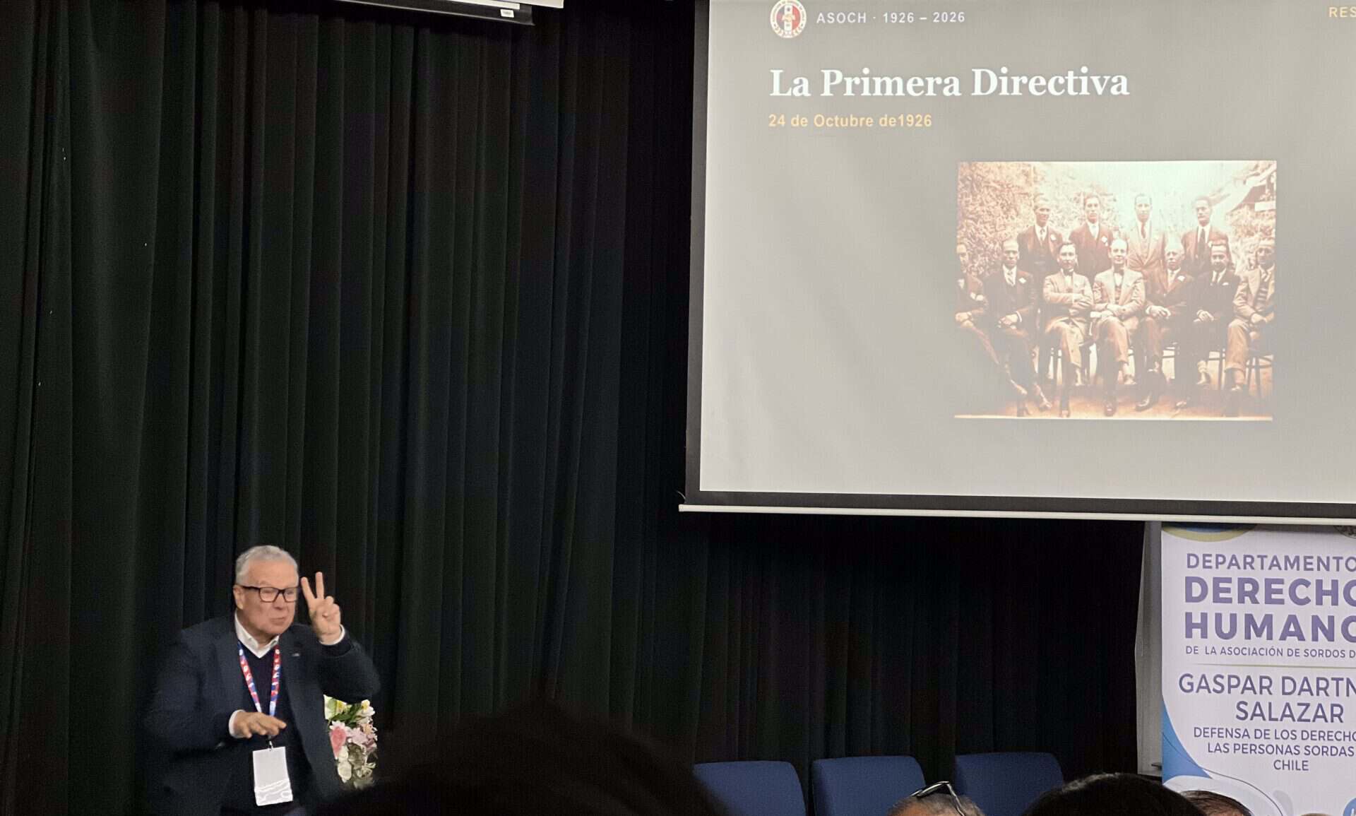 A man stands before a screen showing a historical photo titled “La Primera Directiva” and the date 24 de Octubre de 1926, whilst signing with his hands.
