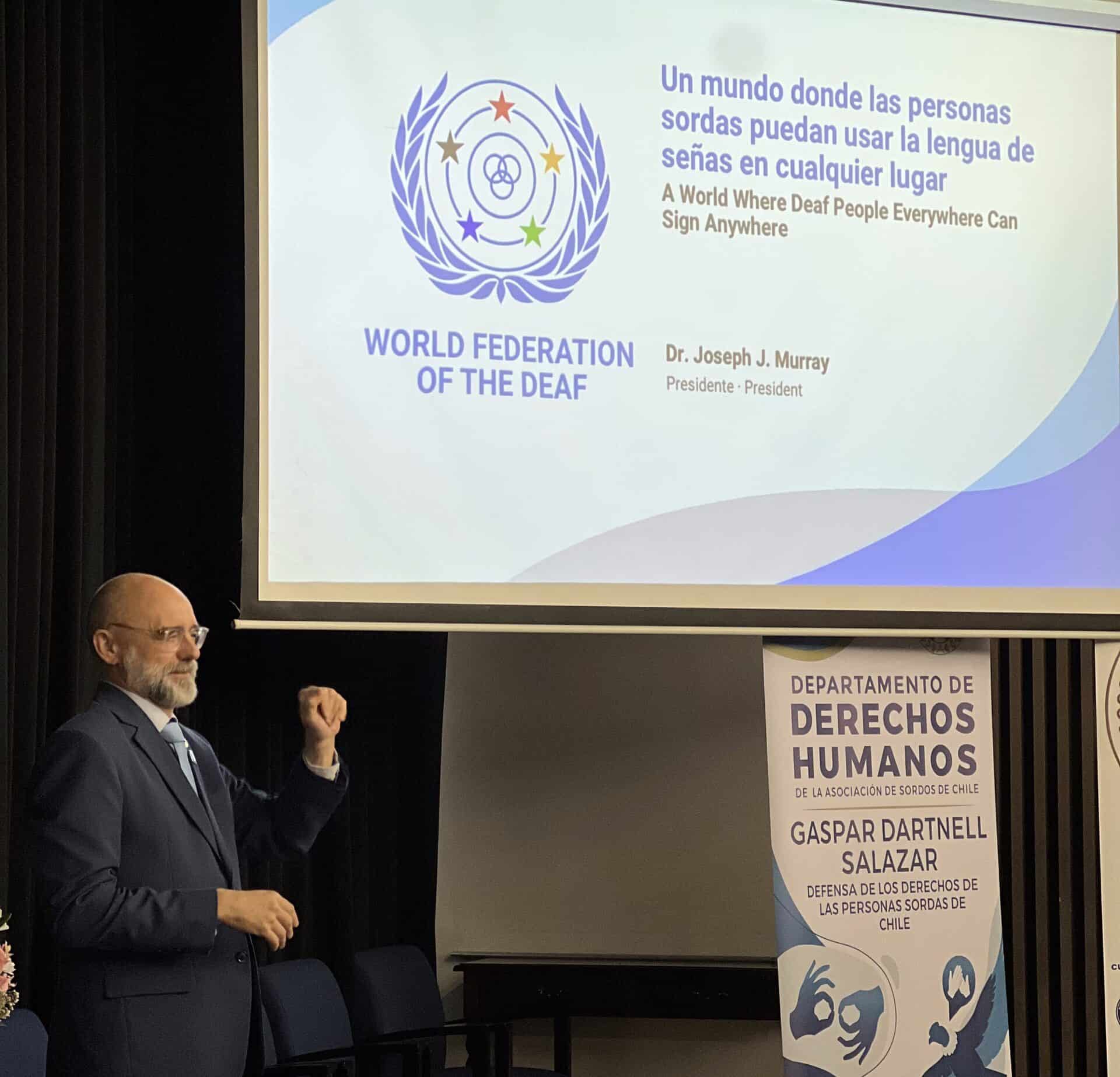 A man in a suit signs in front of a projection screen for the World Federation of the Deaf and a banner for the Human Rights Department of the Association of the Deaf of Chile.
