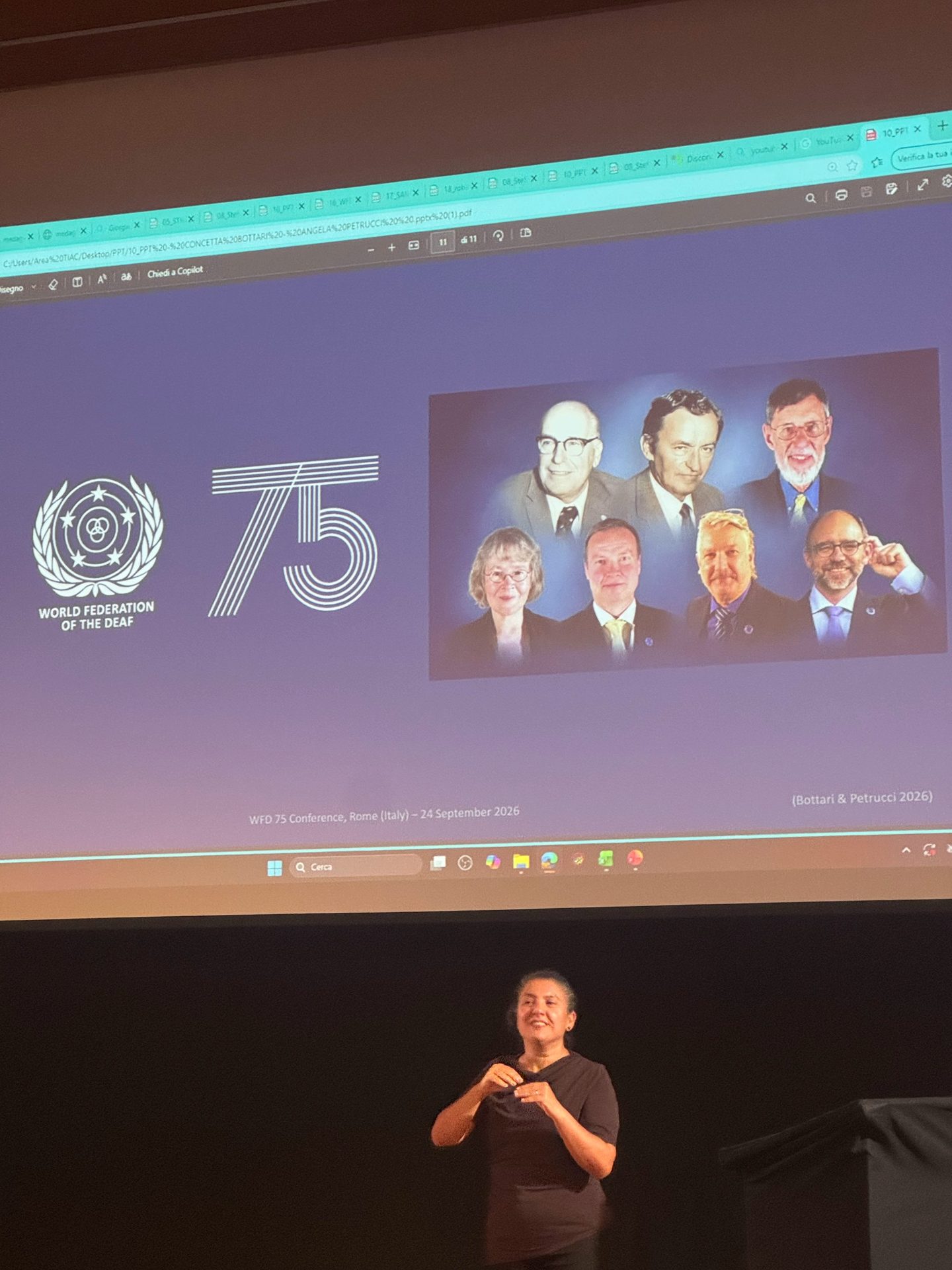 A woman signs on stage in front of a screen displaying the World Federation of the Deaf 75th anniversary logo and portraits of seven people.