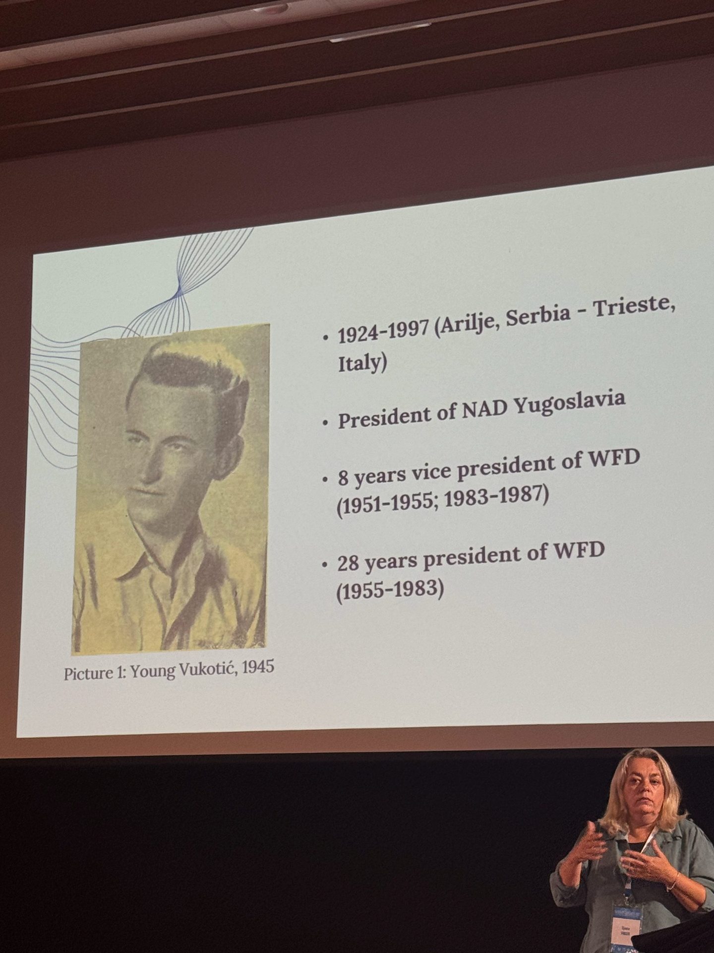 A projected slide shows a black-and-white photo of Young Vukotić (1945) with career highlights; a woman stands in front of the screen presenting.