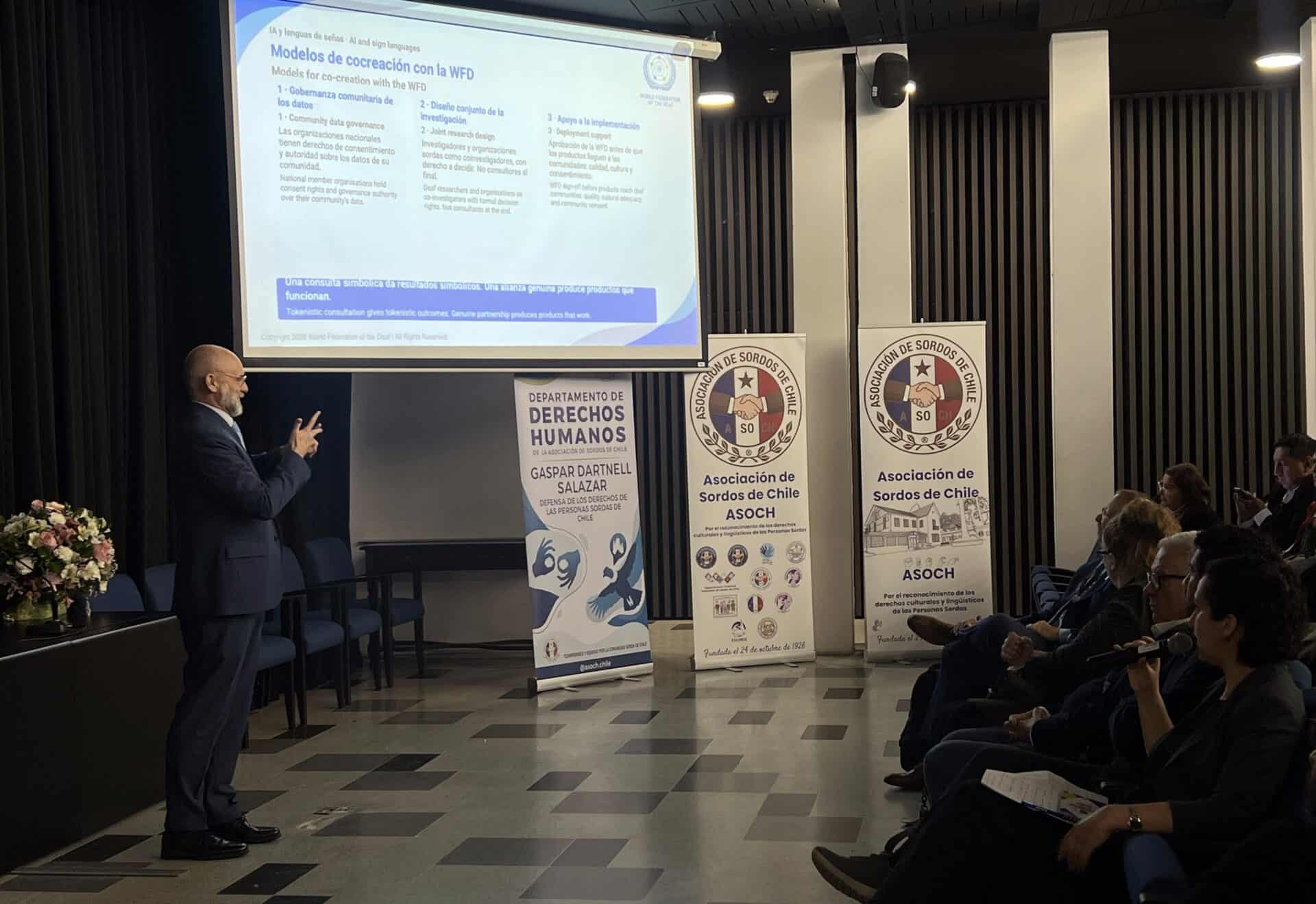 A man in a suit signs to an audience during a presentation on World Federation of the Deaf co-creation models, with ASOCH banners displayed on the stage.