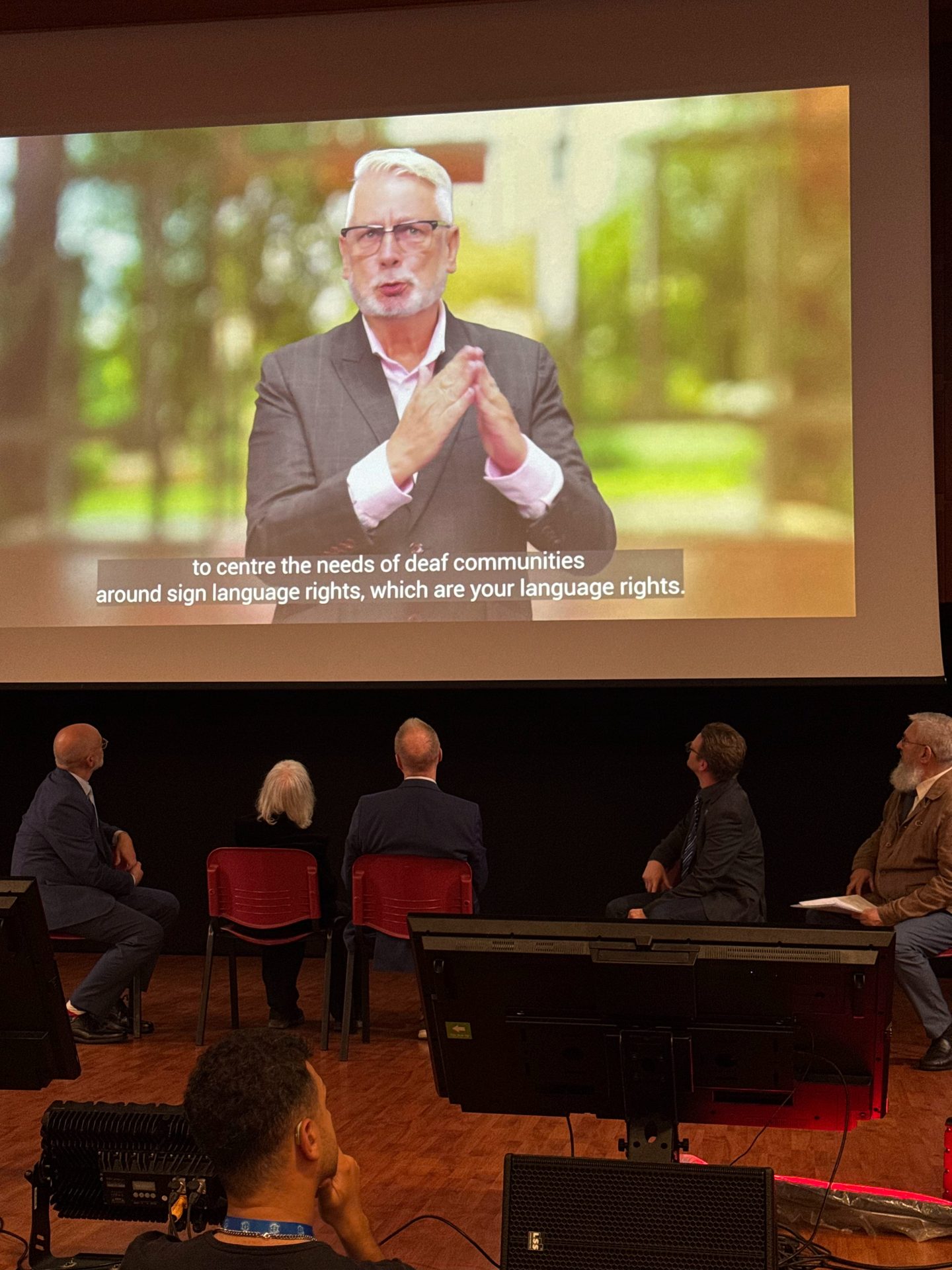 Five people watch a large screen showing a man signing with subtitles about deaf communities and sign language rights.