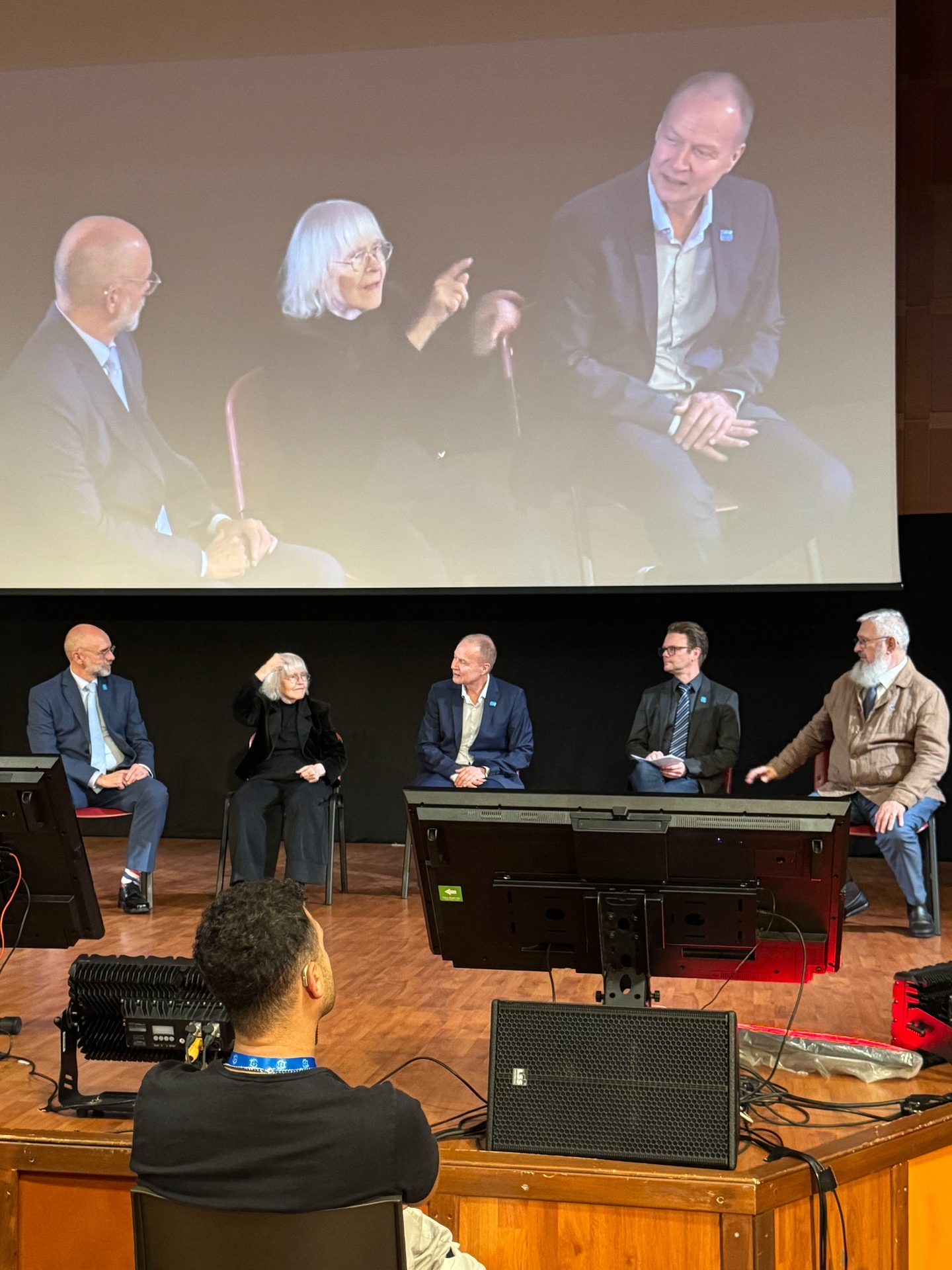 Five people sit on stage in a panel discussion with their image projected on a large screen behind them. An audience member sits in the foreground facing the stage.