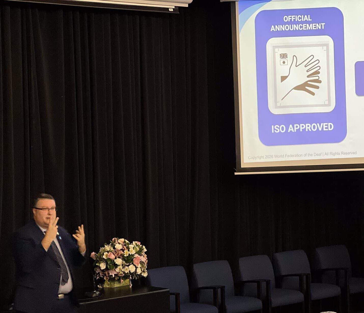 A man signing in front of a projection screen displaying an “Official Announcement” of ISO approval for sign language symbols, alongside a sign for the World Federation of the Deaf human rights department.