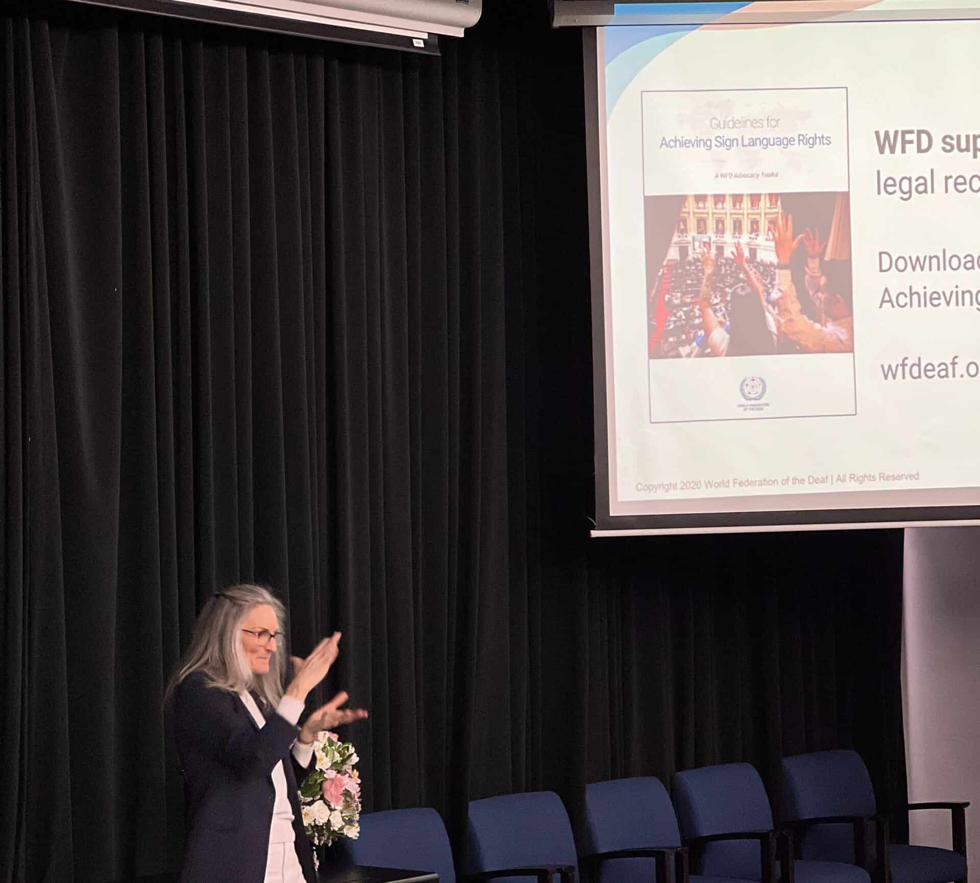 A woman signs in front of a projection screen displaying “Guidelines for Achieving Sign Language Rights”.