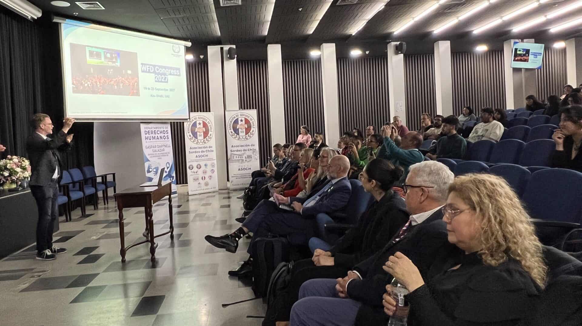 A speaker stands at a lectern in a conference hall addressing an audience seated in rows of blue chairs, with a projection screen displaying “WFD Congress 2027” and banners for the Asociación de Sordos de Chile.