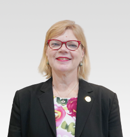 A woman with blonde hair and red glasses, wearing a black blazer over a floral top, poses against a plain light background.