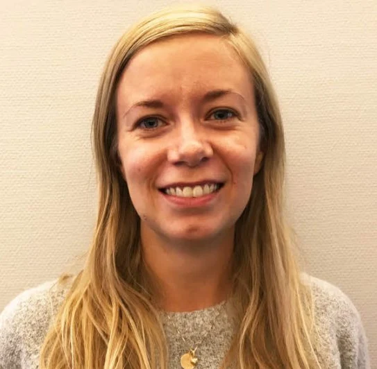 A person with long blonde hair and a gray sweater smiles against a plain background.