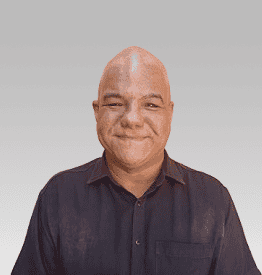 A person with a shaved head is smiling, wearing a dark button-up shirt against a light gray background.