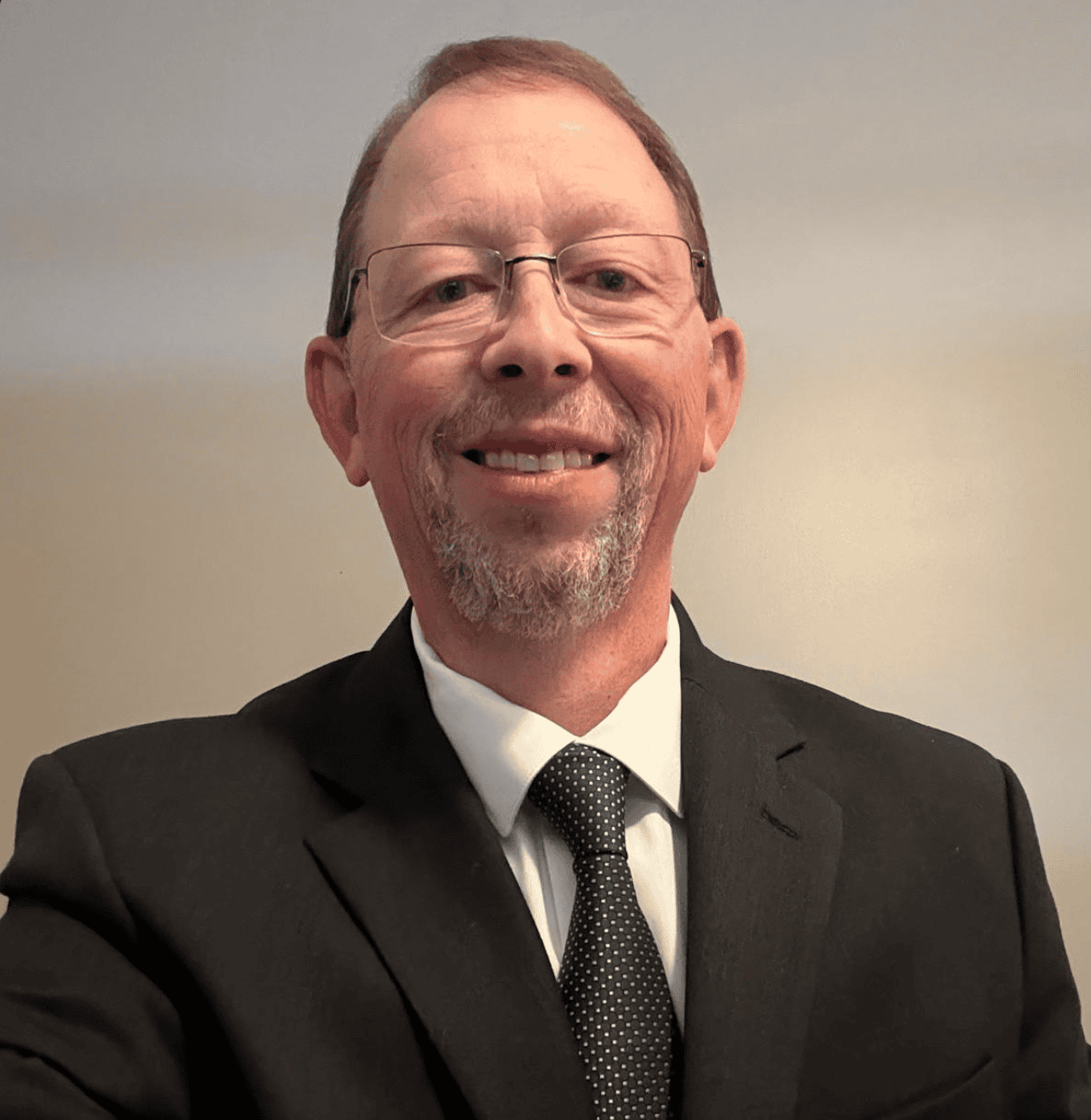 John Moore, WFD Executive Director, smiles at the camera in a black suit, white dress shirt, and dotted black tie, his glasses completing the look against a plain background.