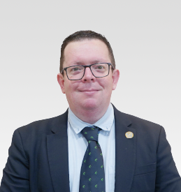 A man wearing glasses, a suit, and a patterned tie is facing the camera and smiling against a plain light background.