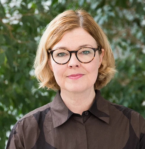 Person with short blonde hair and glasses, wearing a dark shirt, stands in front of greenery.