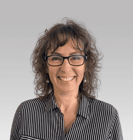 A woman with curly hair and glasses is smiling, wearing a black-and-white striped shirt, standing in front of a plain gray background.