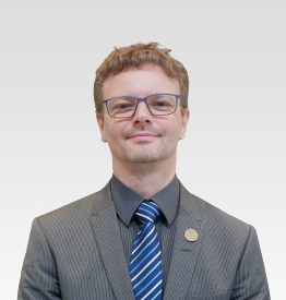 A man in a suit and striped tie stands against a plain white background.