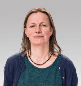 A person with shoulder-length brown hair wearing a teal shirt and navy sweater stands against a gray background.