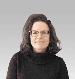 A woman with shoulder-length dark hair and glasses is wearing a black turtleneck sweater against a gray background.