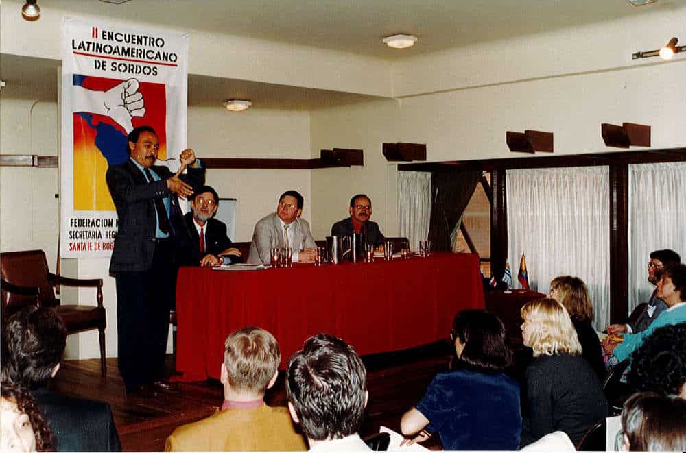 A man speaks at a podium in front of a seated audience during a conference. Three men are seated at a table on stage. A banner on the wall reads "II Encuentro Latinoamericano de Sordos.