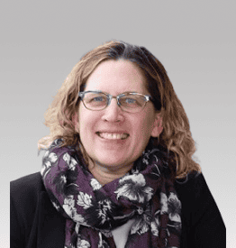 A person with curly hair and glasses is smiling, wearing a patterned scarf and dark jacket against a light gray background.