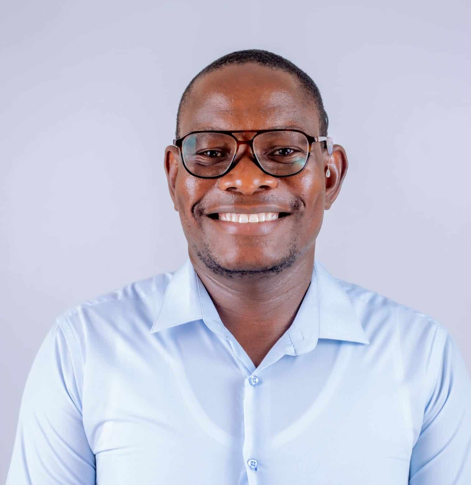Man wearing glasses and a light blue collared shirt smiles at the camera against a plain light background.