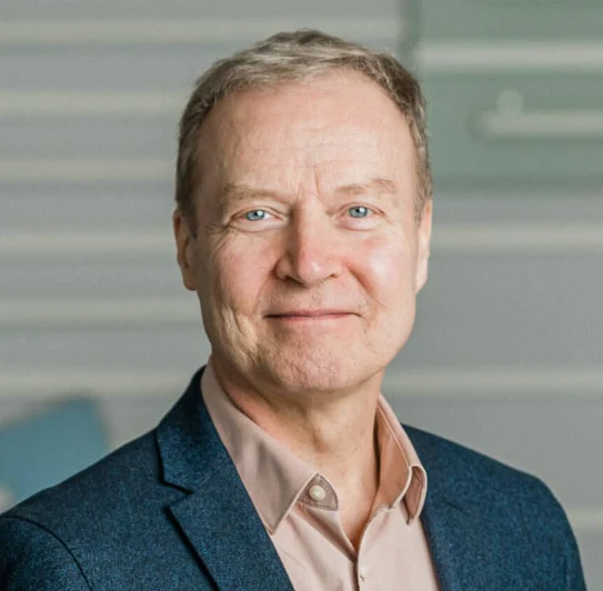 Smiling man in a blazer and shirt with a neutral background.
