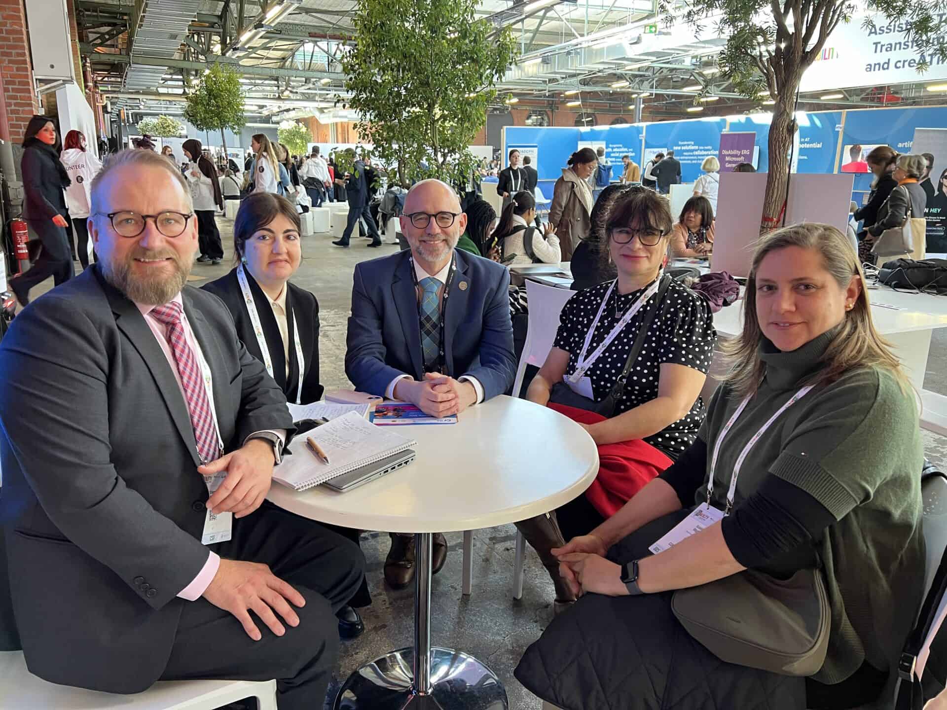 A group of five people sitting around a table at an indoor event, with notebooks and a tree in the background.